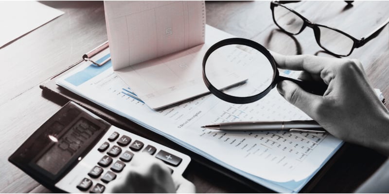 An auditor scrutinises numbers on a balance sheet with a magnifying glass. The time and cost related to auditing has been rising in recent years.